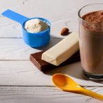 scoop of protein powder an orange plastic spoon and two protein bars placed on top of a wooden table