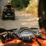 Two ATVs driving on a dirt path with one rider visible in the foreground.