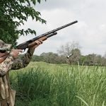 A hunter wearing camouflage gear aiming a shotgun at a flying target in a grassy field.