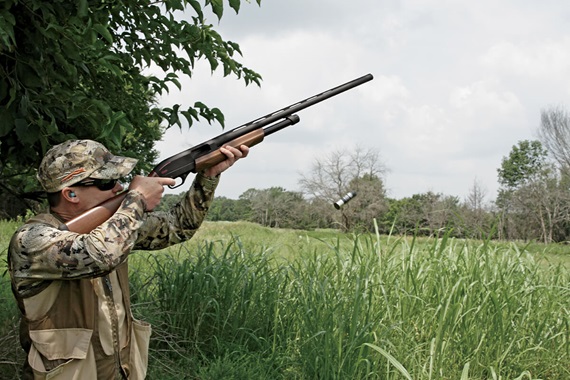 A hunter wearing camouflage gear aiming a shotgun at a flying target in a grassy field.