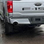 The rear view of a silver Ford F-150 truck, focusing on the tailgate, taillights, and dual exhaust system. The truck is wet from rain, and the photo emphasizes the clean and sleek design of the exhaust tips integrated into the rear bumper.