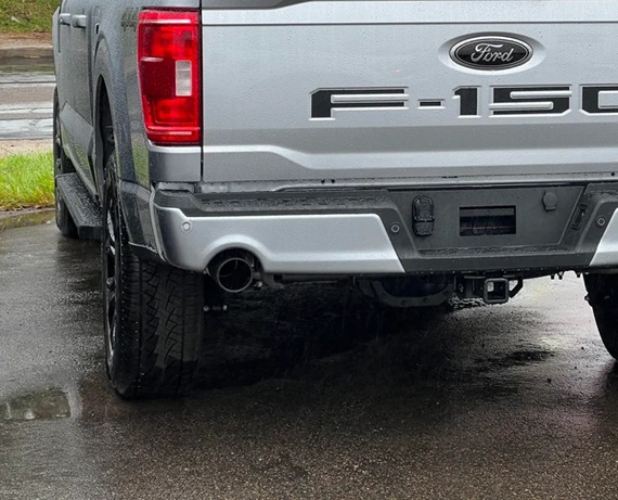 The rear view of a silver Ford F-150 truck, focusing on the tailgate, taillights, and dual exhaust system. The truck is wet from rain, and the photo emphasizes the clean and sleek design of the exhaust tips integrated into the rear bumper.